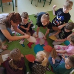 Dzieci siedzą na dywanie i w skupieniu oglądają instrukcję szycia pokazywaną przez nauczycielkę..jpg